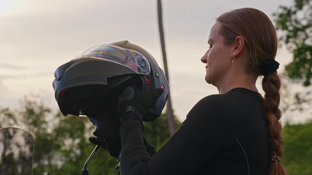 Female Motorcyclist Puts On Black Full Face Helmet and Adjusts Chin Area Outdoors, Protective Riding Gear Supports Road Safety and Injury Prevention