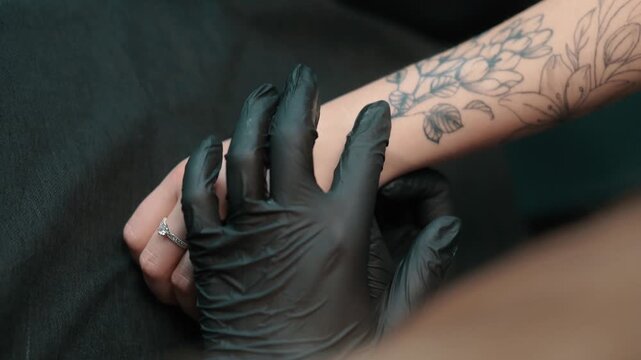 A tattoo artist in black gloves positions a floral stencil on a clients tattooed forearm during a closeup preparation process.