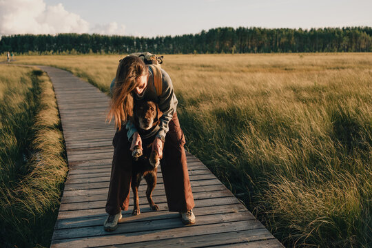 Woman gently holding her dog's front paws on a wooden boardwalk in open countryside, showing pet companionship, human-animal bond, affection and caring outdoor lifestyle.