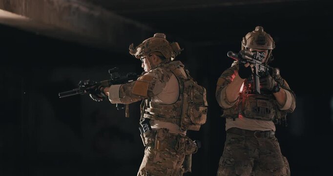 Two tactical operators in camouflage uniforms stand back to back and cover opposite directions with rifles and weapon lights inside a dark abandoned building during a night operation.
