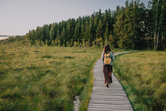 Woman with a backpack walking alone along a wooden boardwalk through open wetlands, illustrating slow travel, mindful outdoor recreation, nature tourism and personal journey.