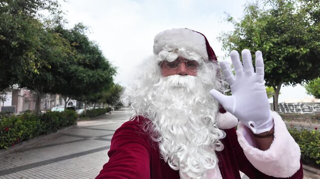 Santa Claus standing outdoors on an urban street, waving and blowing a kiss toward a smartphone camera during a cheerful Christmas video call.