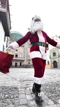 Santa Claus wearing a traditional red and white costume jumping joyfully outdoors while carrying shopping bags filled with Christmas gifts, filmed in slow motion.