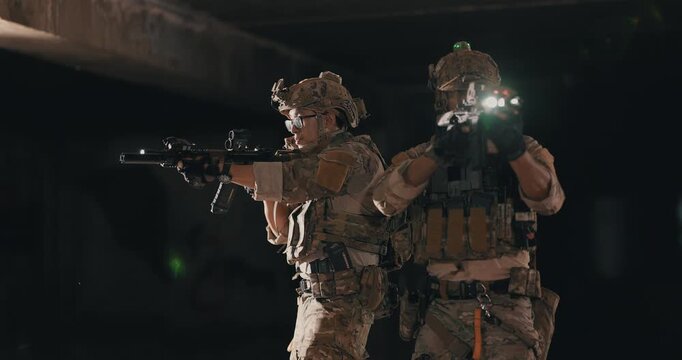 Two tactical operators in camouflage uniforms stand back to back and cover opposite directions with rifles and weapon lights inside a dark abandoned building during a night operation.
