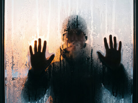 Silhouette of a person with hands pressed against a foggy wet window at sunset