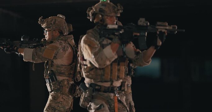 Three  tactical operators in camouflage uniforms stand back to back and cover opposite directions with rifles and weapon lights inside a dark abandoned building during a night operation.
