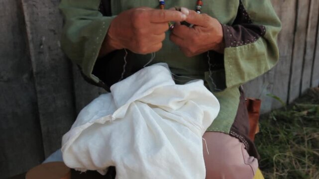 Close up of historical reenactor in authentic medieval clothing hand sewing white fabric with needle and thread