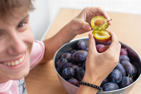 Smiling young man hands breaking a fresh ripe purple plum with a pit over a big metal bowl filled with sweet plums selective focus close up.