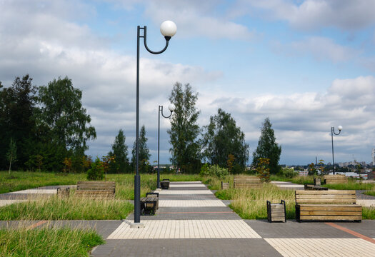 Modern street lamps rise above paved pedestrian walkways and wooden benches in green city park under cloudy sky with copy space