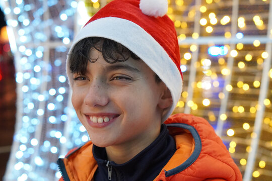 A smiling child wearing a Santa hat in front of some out-of-focus Christmas lights in shades of white and gold