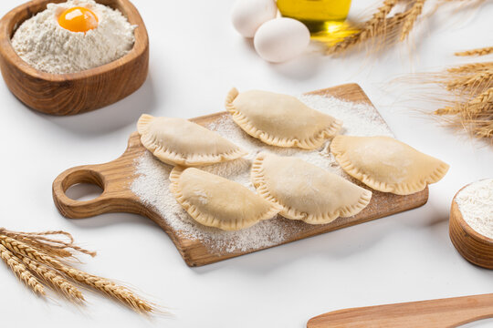 Raw uncooked mini chebureki meat pastries placed on wooden cutting board with flour and eggs on light table