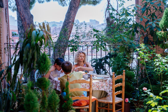 Single mother and two sons sharing quiet breakfast surrounded by plants on home terrace in Spain
