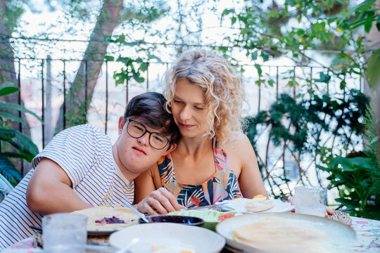 Teenage son with Down syndrome leaning on his single mother during quiet family breakfast in Spain