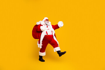 Naklejka premium Santa Claus in red suit carrying a gift sack and walking joyfully on bright yellow background, festive Christmas portrait celebrating holiday cheer and gift giving