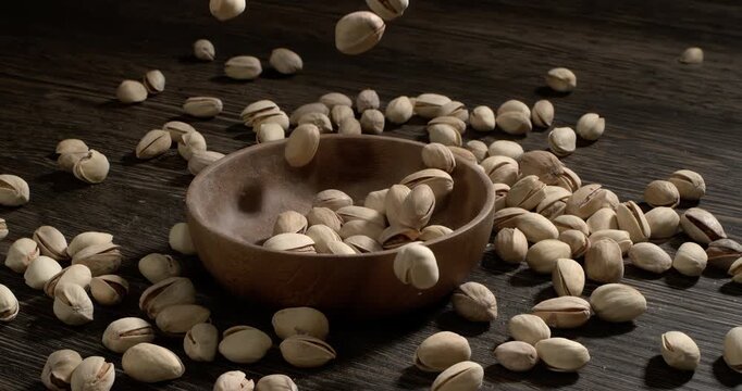 Wooden bowl filled with pistachios on dark wooden table, tasty salted nuts scattered around, healthy snack and natural food concept. Shot on super slow motion camera 1600 fps.