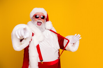 Naklejka premium Santa Claus in red suit and sunglasses pointing and laughing on bright yellow background — festive Christmas portrait with white beard, gloves and suspenders