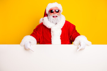Naklejka premium Cheerful Santa Claus in red suit and sunglasses pointing at blank sign on bright yellow background for Christmas holiday promotion and festive advertising