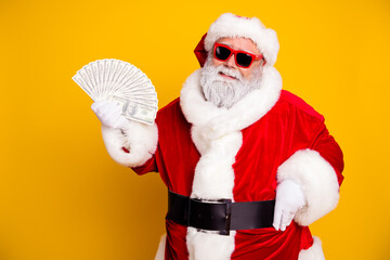 Naklejka premium Santa Claus in sunglasses holding a fan of cash on yellow background, festive Christmas money and holiday wealth concept