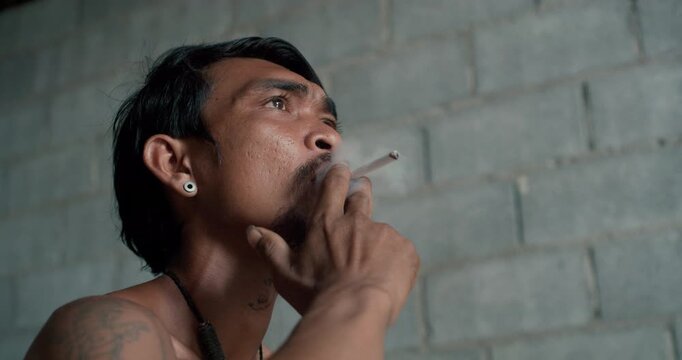 Tattooed man with earring smokes a cigarette, gazing upward against a weathered concrete wall in dim, introspective light.