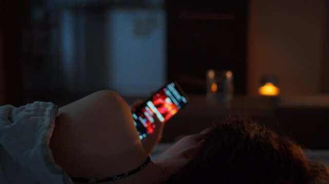 Unable to put her phone down a young woman scrolls through social media in bed late at night. Glowing screen in the dark bedroom. Concept of digital addiction and technology disturbing sleep