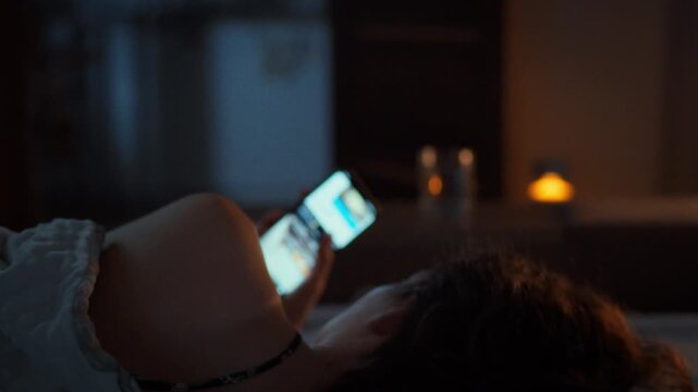 FOMO at night - a young woman lies alone in bed scrolling through social media on her smartphone instead of sleeping. Viewed from behind. Cold dark bedroom. Theme of modern digital habits