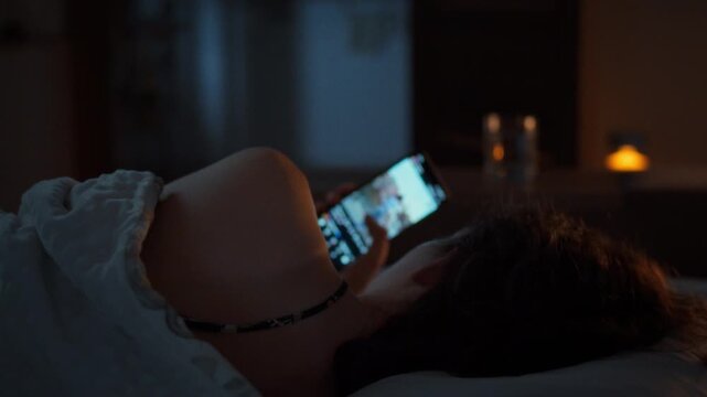 Late at night a young woman cannot sleep and scrolls through social media on her smartphone under a blanket. Dark bedroom. Idea of device dependency and poor sleep in the digital age