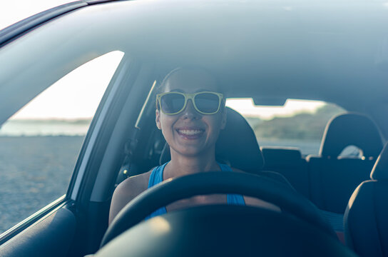A smiling woman wearing yellow sunglasses sits in the driver seat of a car during a sunny road trip.
