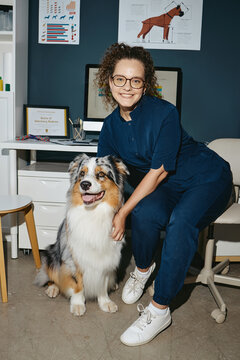 Vertical portrait of young adult Caucasian woman veterinarian smiling while posing with Australian Shepherd dog in vet clinic. Useful for veterinary healthcare promotions