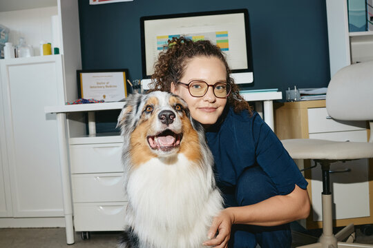 Portrait of Caucasian young adult female veterinarian posing with Australian Shepherd dog in vet clinic office. Image suitable for pet healthcare advertising and veterinary clinic promo