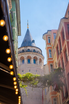 Galata Tower visible between colorful buildings in Istanbul, Turkey, with clear blue sky and decorative lights hanging above the street