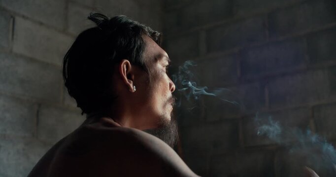 Tattooed man with earring smokes a cigarette, gazing upward against a weathered concrete wall in dim, introspective light.