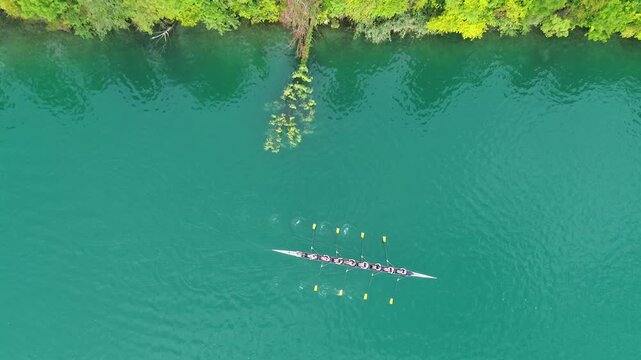 Aerial view of a rowing team training on Lake Bled, Slovenia, with a lakeside road and shoreline partially visible. Turquoise water and reflections create a scenic alpine setting.