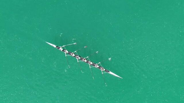 Aerial top-down view of an eight-oared rowing crew training on Lake Bled in Slovenia. Eight rowers move in perfect synchronization across the turquoise water, creating rhythmic patterns and ripples on