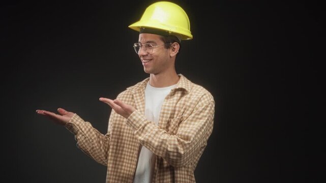 Man wearing a yellow hardhat and clear glasses presents open palms in studio setting under soft lighting; confidence safety helpful.