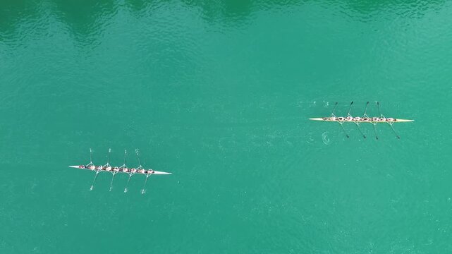Aerial view of two rowing eights training on turquoise Lake Bled, Slovenia. Eight rowers in each crew move in sync, creating reflections and ripples on the water.
