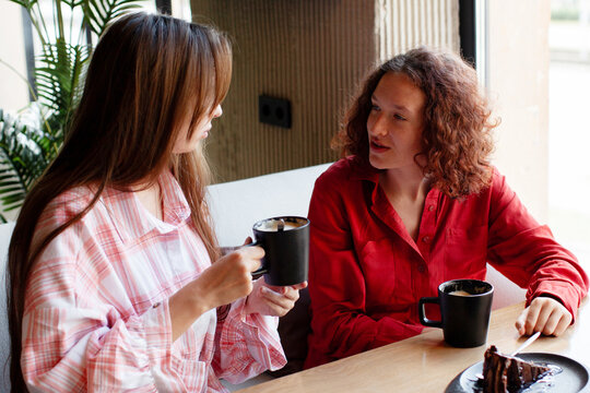Two women talking and drinking coffee together at cafe table. Young friends enjoying conversation with cake in cozy interior. Friendship, communication and leisure time concept.