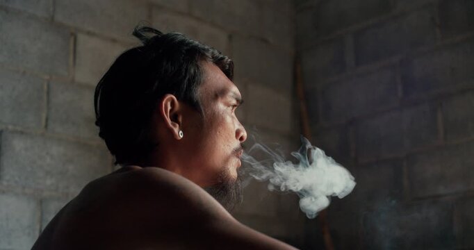Tattooed man with earring smokes a cigarette, gazing upward against a weathered concrete wall in dim, introspective light.