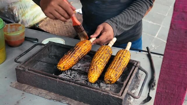 Golden corn cobs sizzle over hot charcoal while a street vendor brushes them with seasoning, turning each cob until the kernels develop a rich, smoky char.