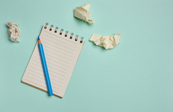 Spiral-bound lined notepad with a blue pencil and crumpled paper balls on a turquoise background.