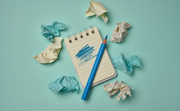 Notepad with a scribbled page and blue pencil surrounded by crumpled paper balls on a turquoise background.