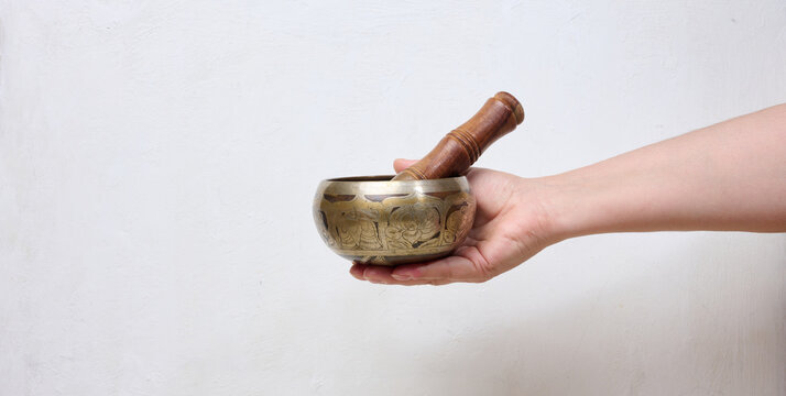 A person holding a metal singing bowl with a wooden stick in their hand against a light background.