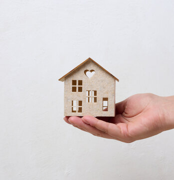 A person holding a small wooden house model on their palm against a light background.