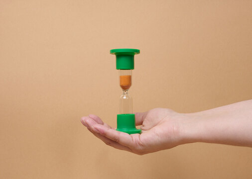 A person holding inverted green hourglass on their palm against a beige background.