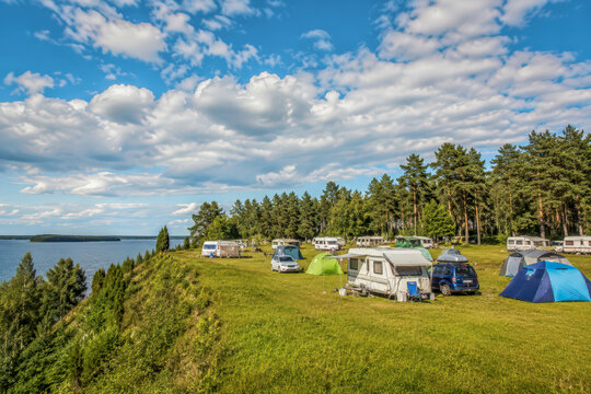 A scenic summer camping site with caravans, tents and cars pitched on a grassy lakeside cliff beside a pine forest under a bright blue sky with puffy clouds.