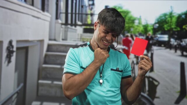 Man referee in turquoise uniform holding red card and clutching throat as if choking while wincing on city street; penalty pain.