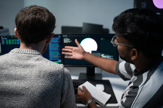 Two colleagues sit together in front of computer monitors while one points at the coding screen to explain software data. They collaborate on a project, sharing ideas and working as a team.