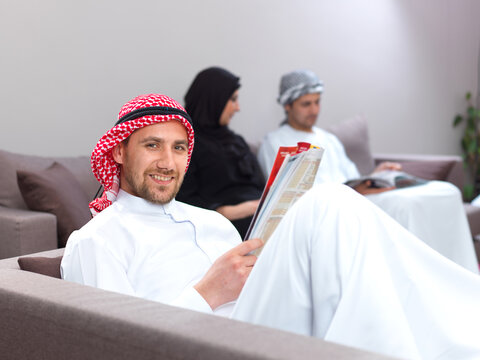 Smiling Arab man holding magazine sitting on couch with people sitting in background in waiting lounge.