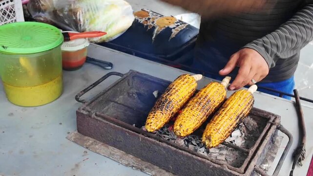 Fresh corn cobs slowly roast over glowing charcoal as a street vendor turns them by hand, creating a smoky aroma and beautifully charred golden kernels ready to serve.