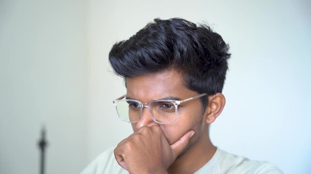 Young man wearing clear eyeglasses sitting indoors with his hand near his face, appearing deep in thought.