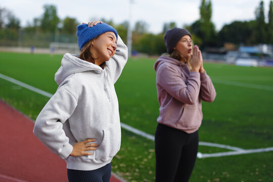 Two women athletes wearing hoodies and beanies preparing for outdoor training on a running track during cold weather, stretching and warming up for their fitness workout